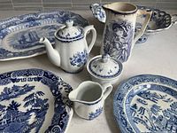 Photo showing entire lot of blue and white china on countertop with visible broken handle on large footed dish, smaller plates, teapot, creamer, sugar bowl and tall pitcher with portrait design.