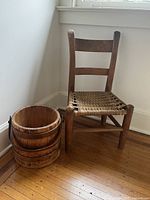 Photo of antique child's chair with woven seat and barrel-style wooden bucket on wooden floor near a white wall.