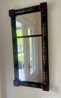 Full front view of the black wooden frame mirror showing gold floral designs, column-style vertical frame sides, and rectangular shape.