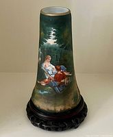 Front view of tall hand-painted porcelain vase showing courting couple under forest trees on a dark green background with wooden base.