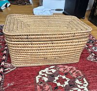 Side view of rectangular antique woven straw bassinet on patterned rug, showing detailed weaving and lid.
