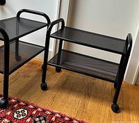 Photo showing both black metal 2-tier rolling utility carts side by side on wooden floor with visible wheels and tubular handles.
