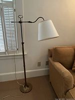 Full view of the bronze articulated floor lamp with a white fabric lampshade positioned next to a tan couch and window with white shutters.