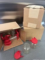 Photo showing open cardboard boxes containing multiple clear plastic jars with red screw-on lids and hinged flaps, with some jars outside the boxes.