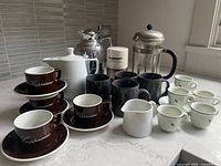 Overall set showing coffee grinder, French press carafe, dark brown cappuccino cups, black mugs, white cups and saucers
