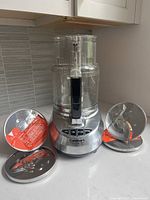 Front view of assembled food processor base with bowl attached and four metal blade discs placed around it on countertop.