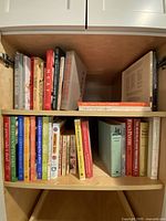 Cookbooks arranged on two shelves inside a wooden cabinet. Books are of various sizes and colors, titles visible including culinary books by various authors.
