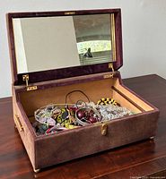 Open faux-suede covered jewelry box showing assorted jewelry inside with interior mirror on lid