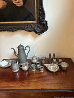 Full view of entire pewter tea set and related pieces arranged on wooden table under a framed painting.