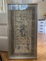 Vertical framed Asian cityscape artwork in silver frame with visible window reflection, wood floor background