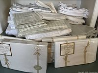 Stack of bed linens with some packaged sheet sets and others loosely folded, showing white and beige tones with some checkered patterns.