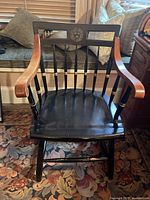 Front view of black Windsor style armchair with brown armrests and Harvard University emblem on backrest, placed on floral carpet near window seat.