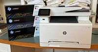 HP Color LaserJet Pro MFP M277dw laser printer with touchscreen and document feeder shown alongside three toner cartridge boxes.