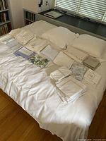 Full shot of king size bedding set laid out on bed including white quilted cover, multiple folded sheets, pillowcases in white and purple, and floral-patterned linens.