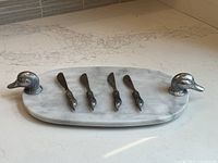 Oval marble cheese tray with metal duck-head handles and 4 duck-handle utensils arranged on marble countertop.