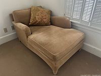 Front angled view of dusty caramel chenille chaise lounge with decorative pillow on a beige carpet, near white shuttered window and white wall.