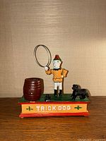 Front view of multicolor cast iron trick dog mechanical coin bank showing the standing figure with hoop, barrel, and dog on base labeled 'TRICK DOG'.