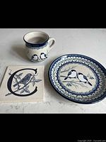 Photo shows cup, round plate with birds and floral pattern, and square trivet with bird and letter C design placed on a countertop.