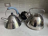 Three tea kettles displayed together on a white countertop under a tiled backsplash: Alessi kettle (left) with blue handle, black ceramic teapot (center back), All-Clad kettle (right) with a large curved handle.