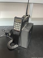 Vintage Radio Shack cell phone from the 1980s-1990s era shown on a black surface. Includes handset with numeric keypad and display, coiled cord, base unit with antenna, and power cord.