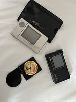 Photo showing three items: silver Citizen 6T-4QA pocket TV with LCD screen and speaker, black Casio TV-200 pocket TV, and black folding Seiko travel clock with gold-trimmed round analog face