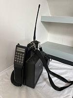 Vintage Radio Shack cellular phone with battery pack, antenna, and strap placed on a white cloth background, showing the full unit.