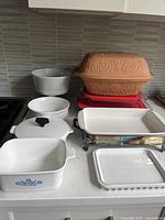 Photo showing the full set of bakeware including the terracotta Romertopf chicken pot at top, white ceramic casseroles with lids, Corning Ware dish with blue pattern, and rectangular baking dishes.