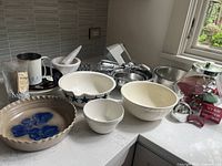 Photo showing assortment of ceramic bowls, metal utensils, hand mixer, and glass jar with cookie cutters on kitchen counter.