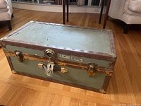 Top and front view of a rectangular vintage steamer trunk with green panels and brown trim, metal corner protectors and central lock mechanism without key