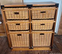 Front view of wooden storage unit with six wicker baskets in two columns of three. Each basket has hand holes for easy handling. Unit has black metal corner reinforcements and natural wood finish.