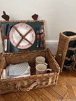 Open wicker picnic basket showing two plates secured with leather straps, two ceramic mugs, a set of cutlery with dark handles, and folded cloth napkins inside.