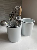 Two white ceramic utensil holders with black rim trim on countertop, one holding various kitchen utensils including stainless steel spoons, a whisk, a wooden spoon, and a slotted spatula.