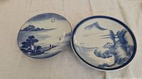 Top view showing two ceramic plates with blue and white landscape scenes, one with a moon and mountains and another with a house and mountain
