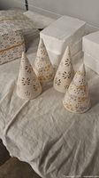 Four white porcelain Christmas trees with gold and silver painted snowflake and star cutout patterns, arranged on a cloth surface.