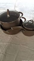 Photo showing large saucepan, smaller saucepan, and skillet all in bronze metal finish with glass lids on the saucepans.