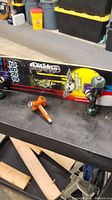 Complete skateboard displayed on a black surface, showing side view with trucks, green wheels, part of graphic deck artwork, and skateboard tool next to it.