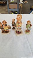 Front view showing all four Hummel figurines of children, including a girl holding a doll, two children with baskets, child with orange hat holding watering can, and child with hand near mouth.