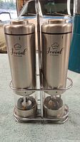 Two Jovial Kitchen branded salt and pepper grinders made of stainless steel and glass in a metal stand on a surface with natural light showing slight signs of use.