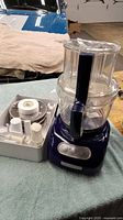 Blue KitchenAid food processor with clear bowl and handle on blue towel, with white plastic attachment tray beside it.