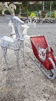 Outdoor photo showing large lighted metal wire reindeer and red and silver sleigh decoration on gravel surface.