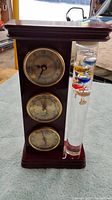 Front view showing the wooden stand with clock, barometer, hygrometer dials and Galileo thermometer tube with colorful spheres and temperature tags.