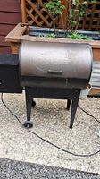 Full view of metal Traeger pellet grill with lid closed and two wheels visible, placed outdoors on gravel and concrete ground.