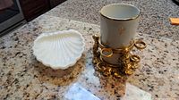 Photo showing top view of white scallop shell soap dish, ceramic cup with gold Fleur-de-lis and ornate gold metal base holding the cup