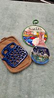 Three Seattle landmarks themed items: a ceramic decorative plate showing city scenes, a tan fabric pot holder, and a blue metal trivet shaped like a kettle.