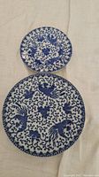 Two Japanese ceramic plates placed separately on a fabric surface, showing blue decorative pattern on white/off-white ceramic and minor wear.