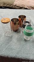Set of five mugs and cups on a soft blue surface including two copper-tone hammered mugs, two glass cups with colored bands, and one clear plastic tumbler with bamboo lid and straw.