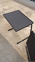 Black folding table with plastic top and foldable metal legs shown on concrete floor.
