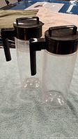 Two clear plastic pitchers with black lids and handles on a cloth surface.