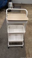 Front view of the three-tier white metal rolling cart with wood-look removable top and two perforated lower shelves.