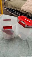 Two containers; one pitcher with handle and red lid, and one separate container with red clip lid, both made of transparent plastic.